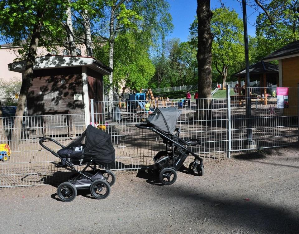 Två barnvagnar står parkerade utanför en inhägnad lekpark.