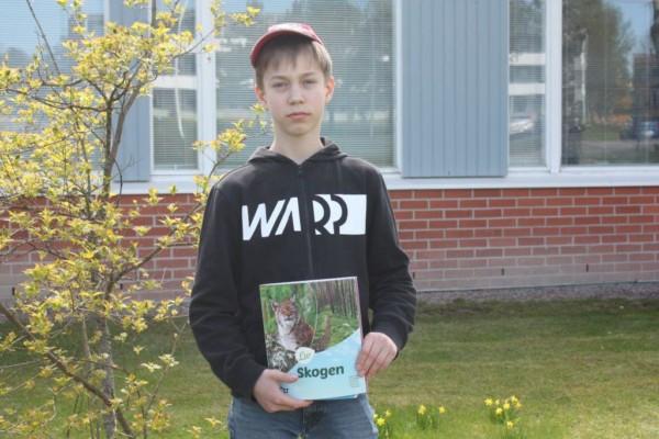 Samuel Engblom med röd keps på huvudet, håller i sin biologi bok med titeln "Skogen", i bakgrunden en buske och skolhuset.