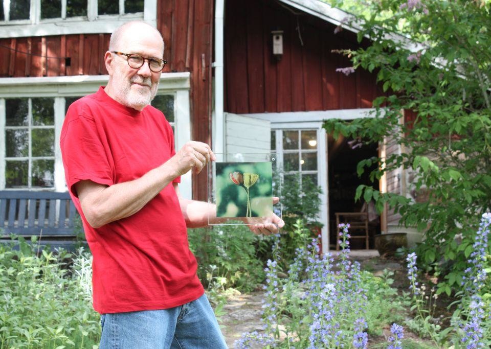 Janne Gröning håller i en glastavla med blomtryck, står utanför det röda huset i Westers Trädgård som han ställer ut i, grönt träd i bakgrunden och lila blommor runt omkring