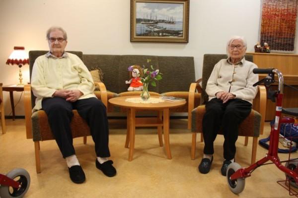 Margit Karlsson och Violet Åhlfors sitter i varsin stol, mellan dem står ett litet runt bord, i bakgrunden en grön soffa och en tavla på väggen