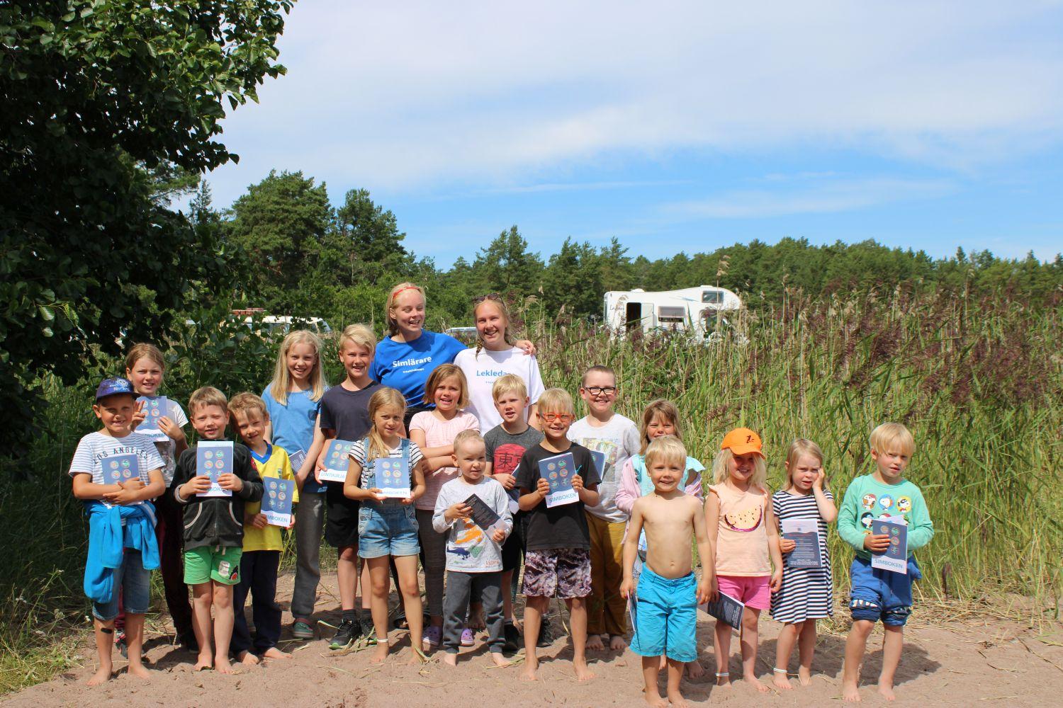 17 barn håller i sina simskolediplom på den soliga sandstranden. Simläraren och lekledaren står bakom dem.