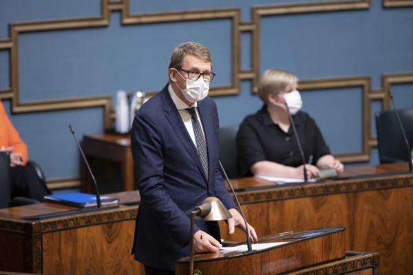 Kostymklädd herre i ansiktsmask vid riksdagens plenisals podium