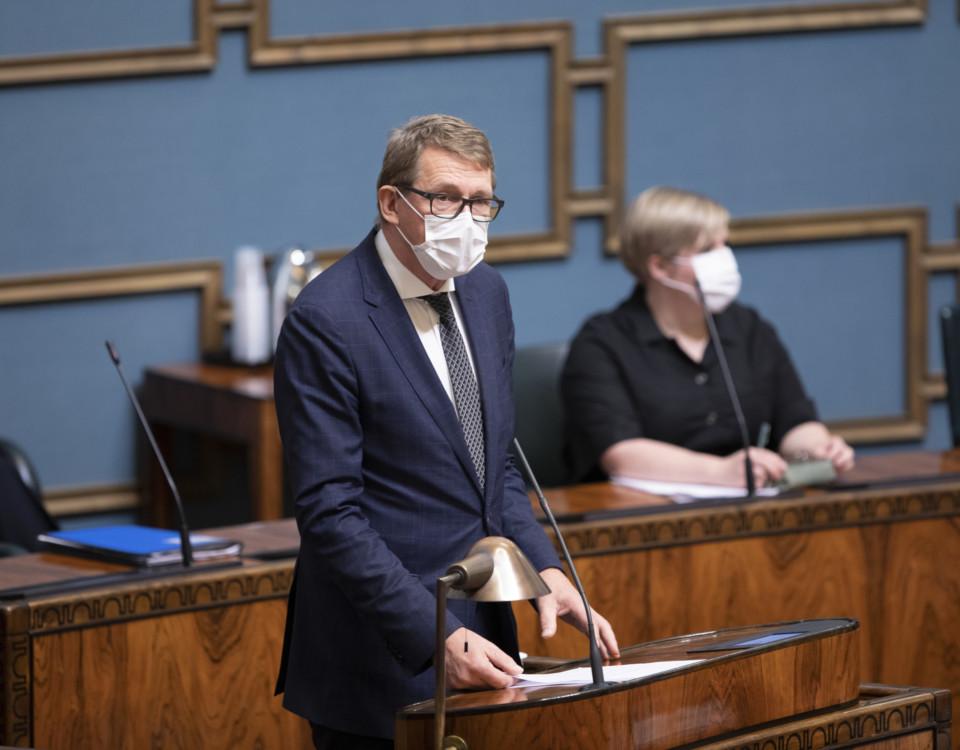 Kostymklädd herre i ansiktsmask vid riksdagens plenisals podium