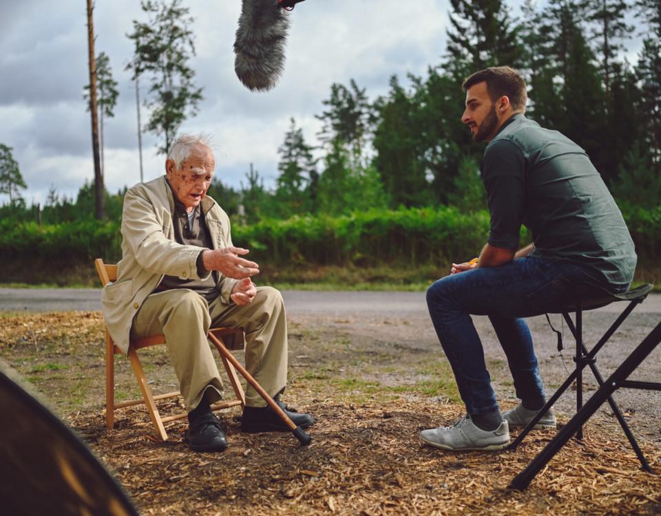 En ung man intervjuar en krigsveteran. De sitter på stolar utomhus, bilden är tagen en sommardag.