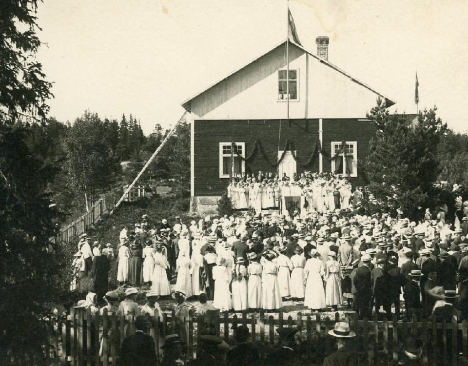 en gammal svartvit bild på ett hus och en mängd festklädda människor på gården