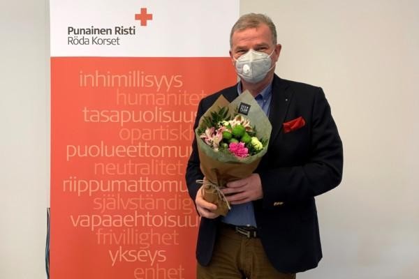 man ikkädd munskydd står och håller i en blombukett. han står framför Röda Kors-väg