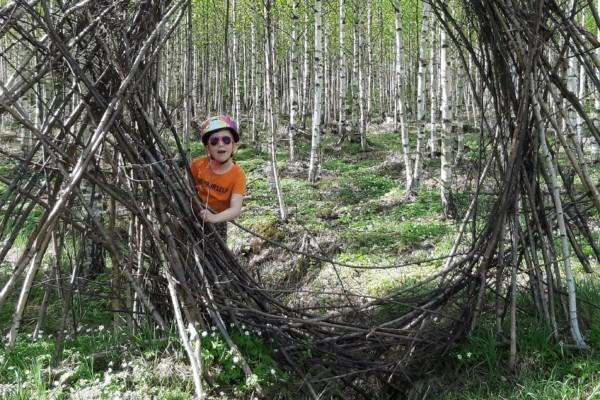 Liten flicka i orange t-tröja, solglasögon och hjälm kikar ut ur en prot gjord av tunna trädstammar.