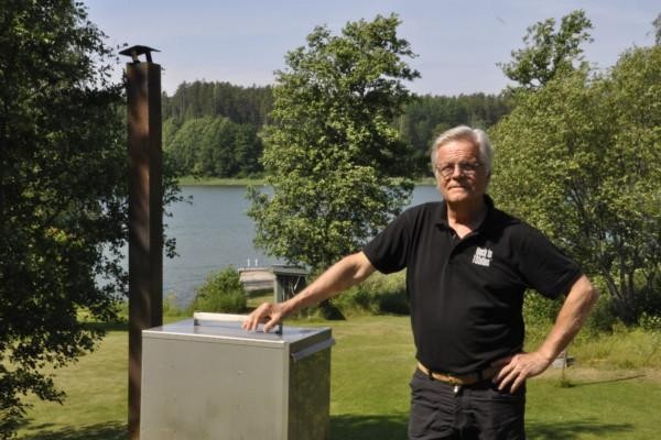 Man i svart pikéskjorta bredvid en rökugn i metall, i bakgrunden gröna träd, gräsmatta och vatten.