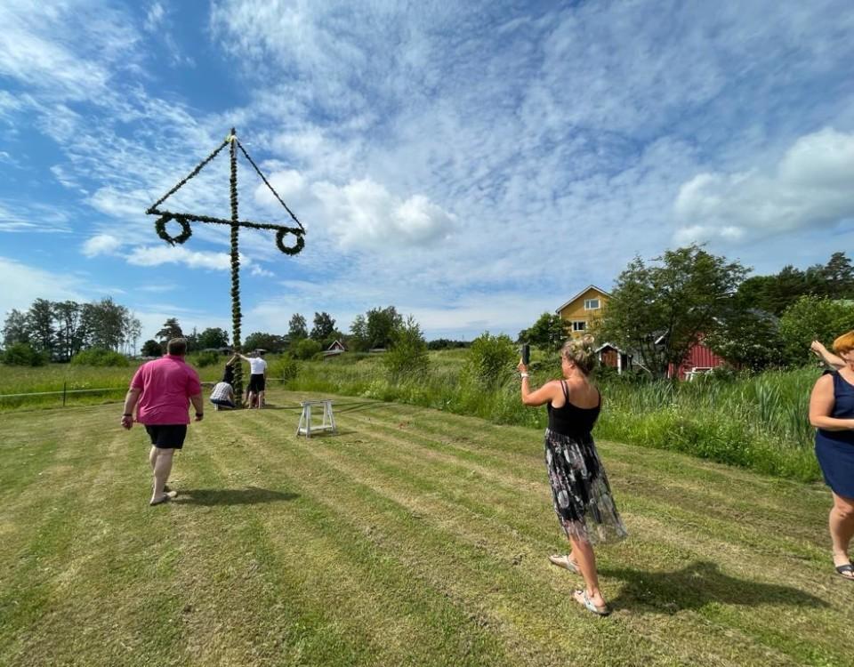 En gräsmatta där en ståtlig midsommarstång står. Enstaka människor syns också på bilden, närmast kameran en kvinna som fotograferar stången.