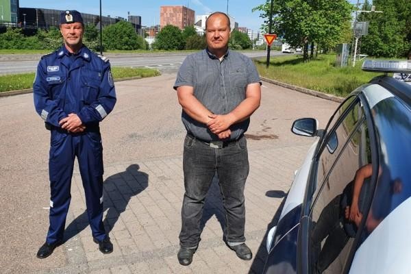 Peltola iklädd polisuniform och Heiskanen i skorta och jeans poserar framför en polisbil med motorvägen i bakgrunden.