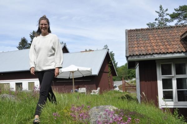 Kvinna i svarta byxor och vit tröja ler mot kameran, röda stugor i bakgrunden.