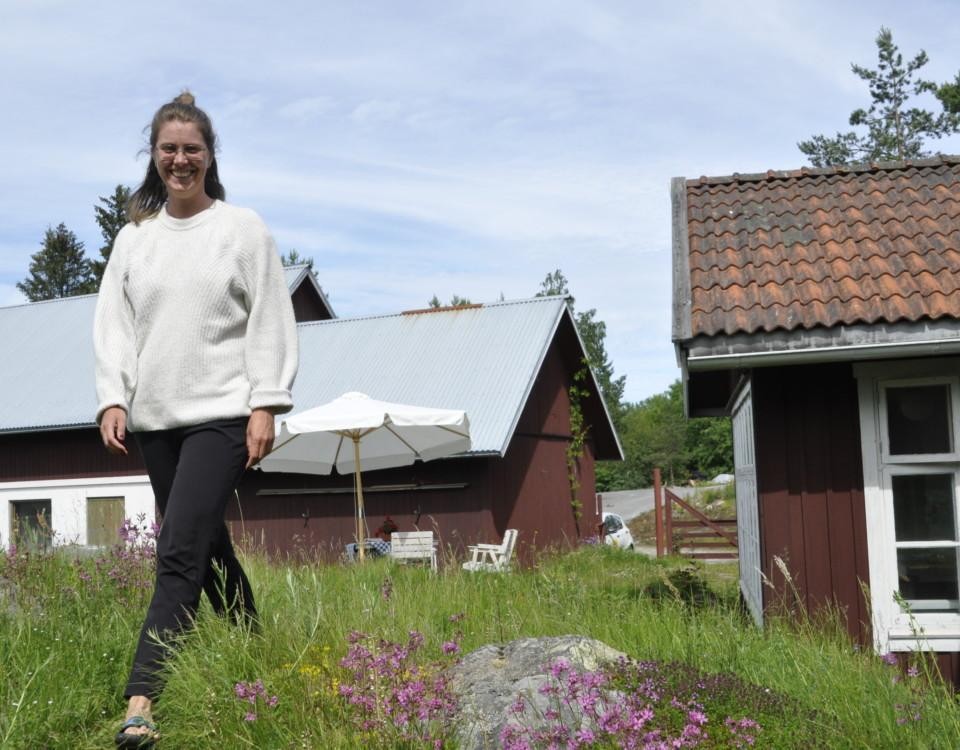 Kvinna i svarta byxor och vit tröja ler mot kameran, röda stugor i bakgrunden.