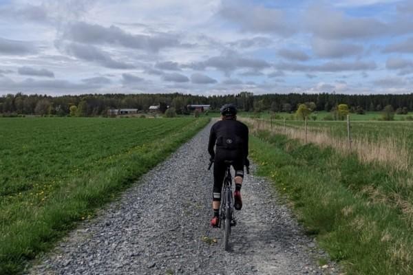 Svartklädd cyklist på grusväg, gröna åkrar på sidor om vägen, skog i bakgrunden.