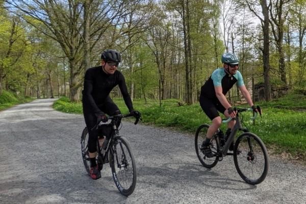 Två män cyklar på varsin cykel på grusväg, träd i bakgrunden