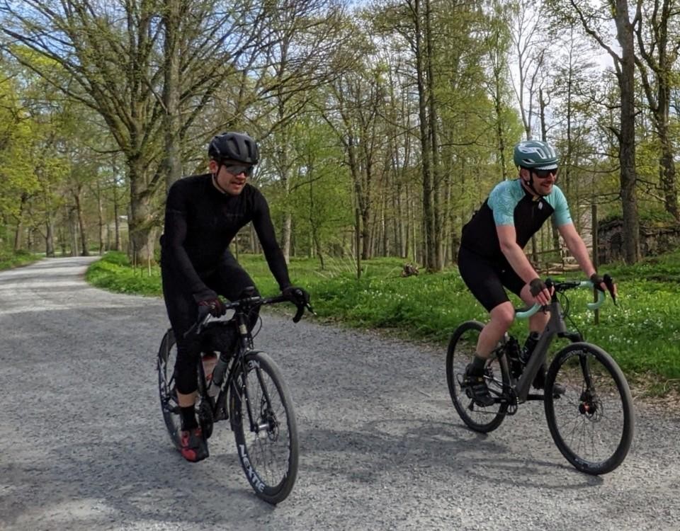 Två män cyklar på varsin cykel på grusväg, träd i bakgrunden