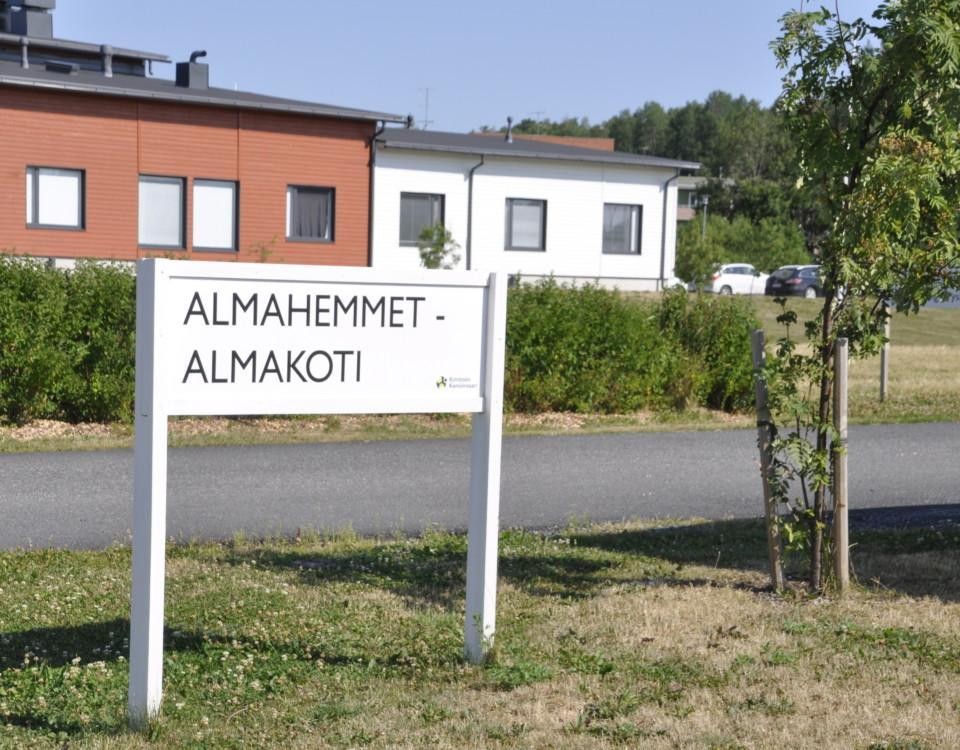Skylt med texten "Almahemmet - Almakoti", rödvitthus i bakgrunden, grönt buskage runt omkring