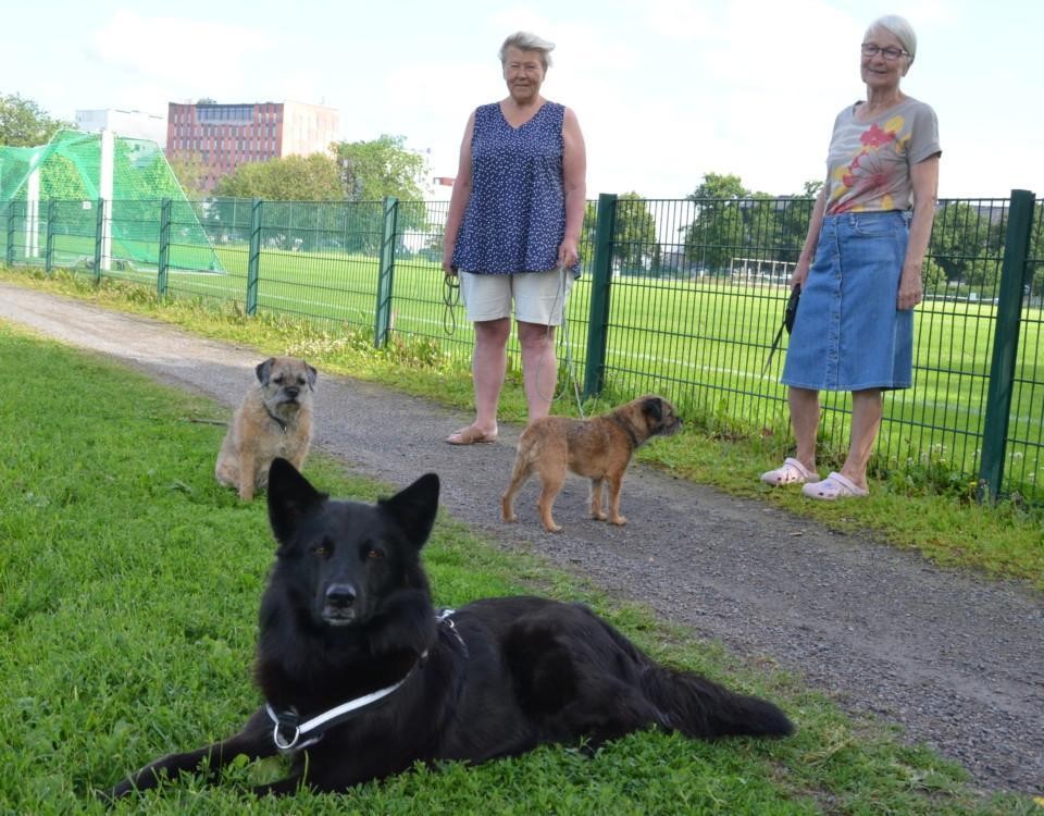 Två damer står på en gräsmatta med sina tre hundar, två små och en större svart