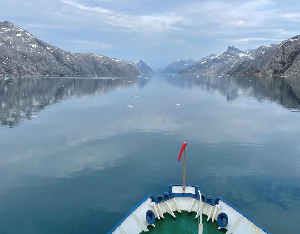 Fören på en båt som färdas i en fjord. Havsytan är glasblank.