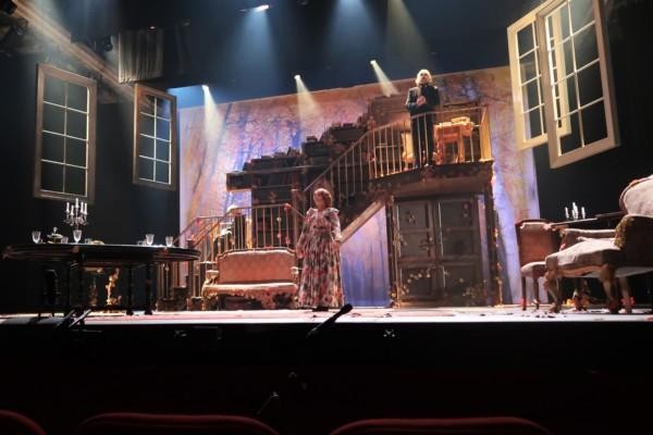 Bild av teaterscen. En kvinna i blommig klänning och en skäggig man på en trappa står på scennen.