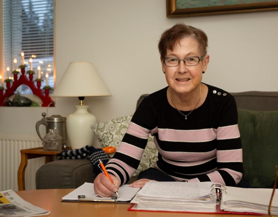 äldre kvinna sitter i soffa med papper och penna framför sig på soffbordet