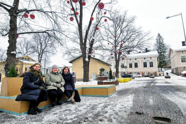 Tre kvinnor på en bänk under träd pyntade med stora röda bollar.