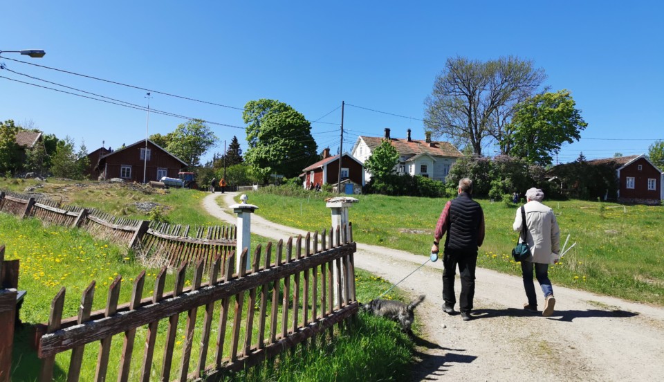 Två män som går med hund på ö med vitt hus i bakgrunden.