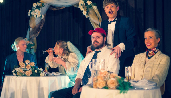 Skådespelar på scen i Skärgårdsteatern. Brudpar i bakgrunden som ser oroliga ut, längst fram brudens far och farmor tillsammans med en ung man som är brudens ex.