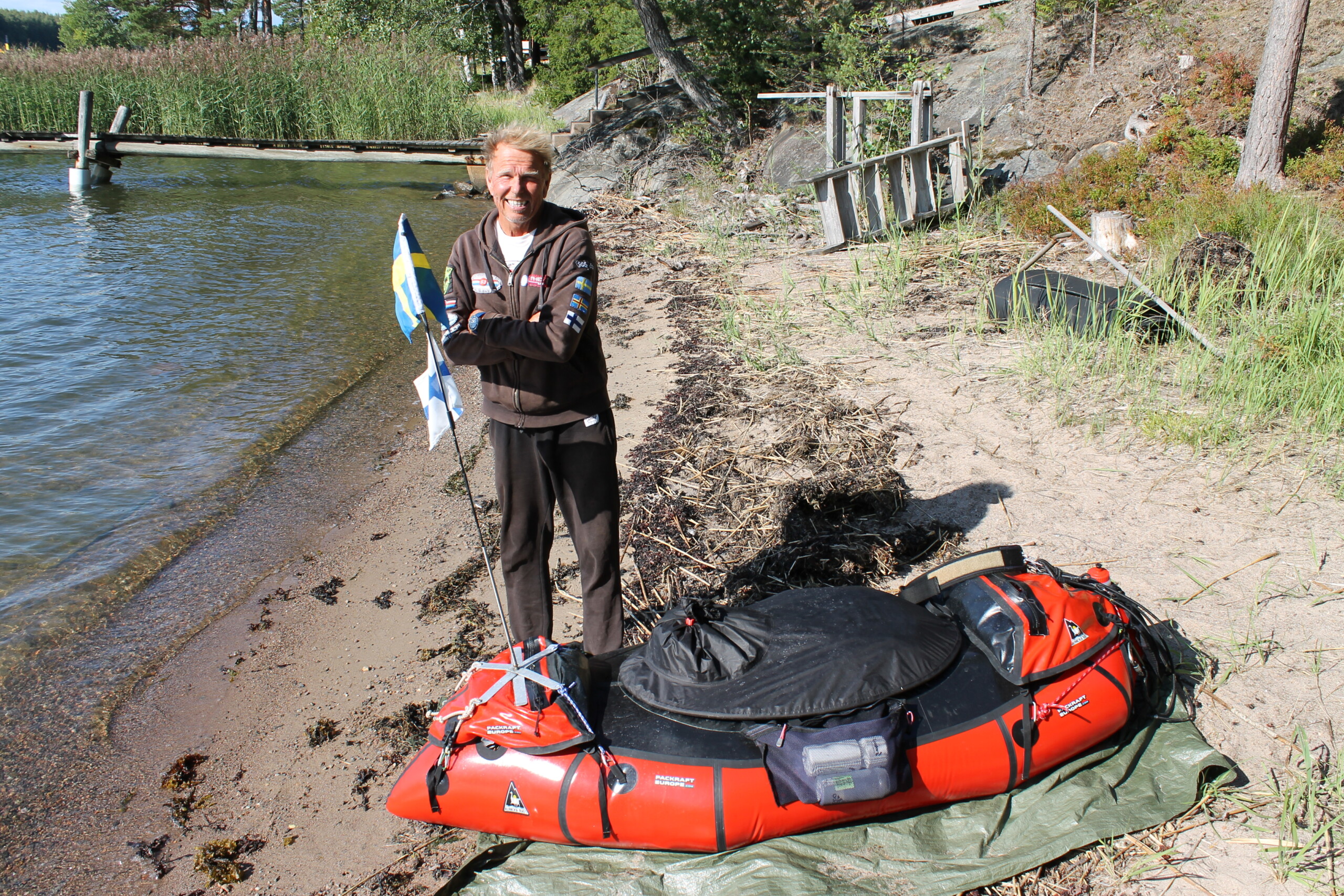 En man står på en strand vid en liten båt. Han har ljust kommit i land med båten han drar efter sig då han simmar.