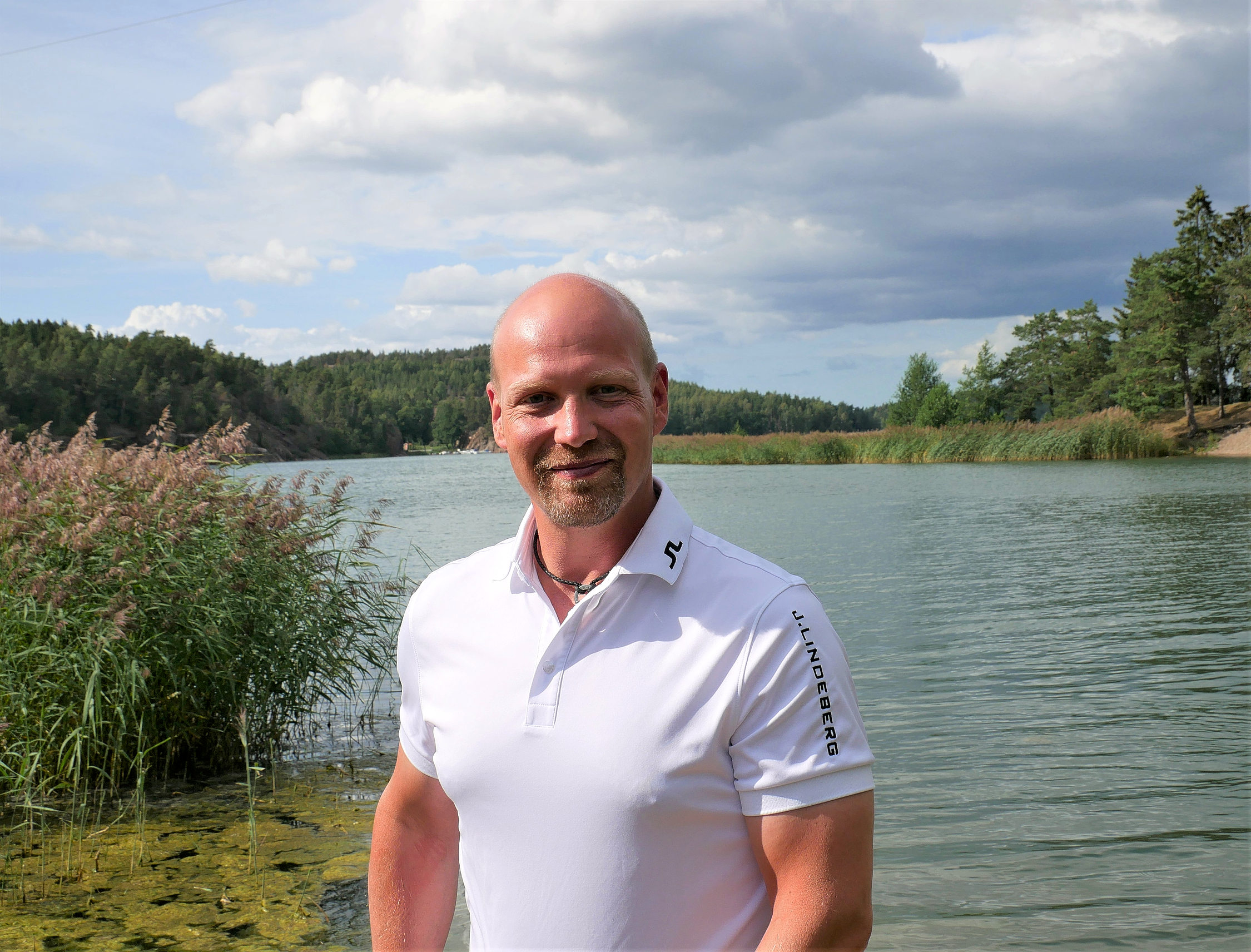 Man i vit skjorta står på stranden med vattnet i bakgrunden