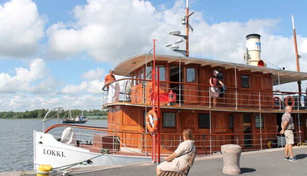 En gammal ångbåt står i en hamn. Vatten och himmel syns i bakgrunden. På båten går flera människor omkring. På en bänk vid båten sitter en kvinna på en bänk.