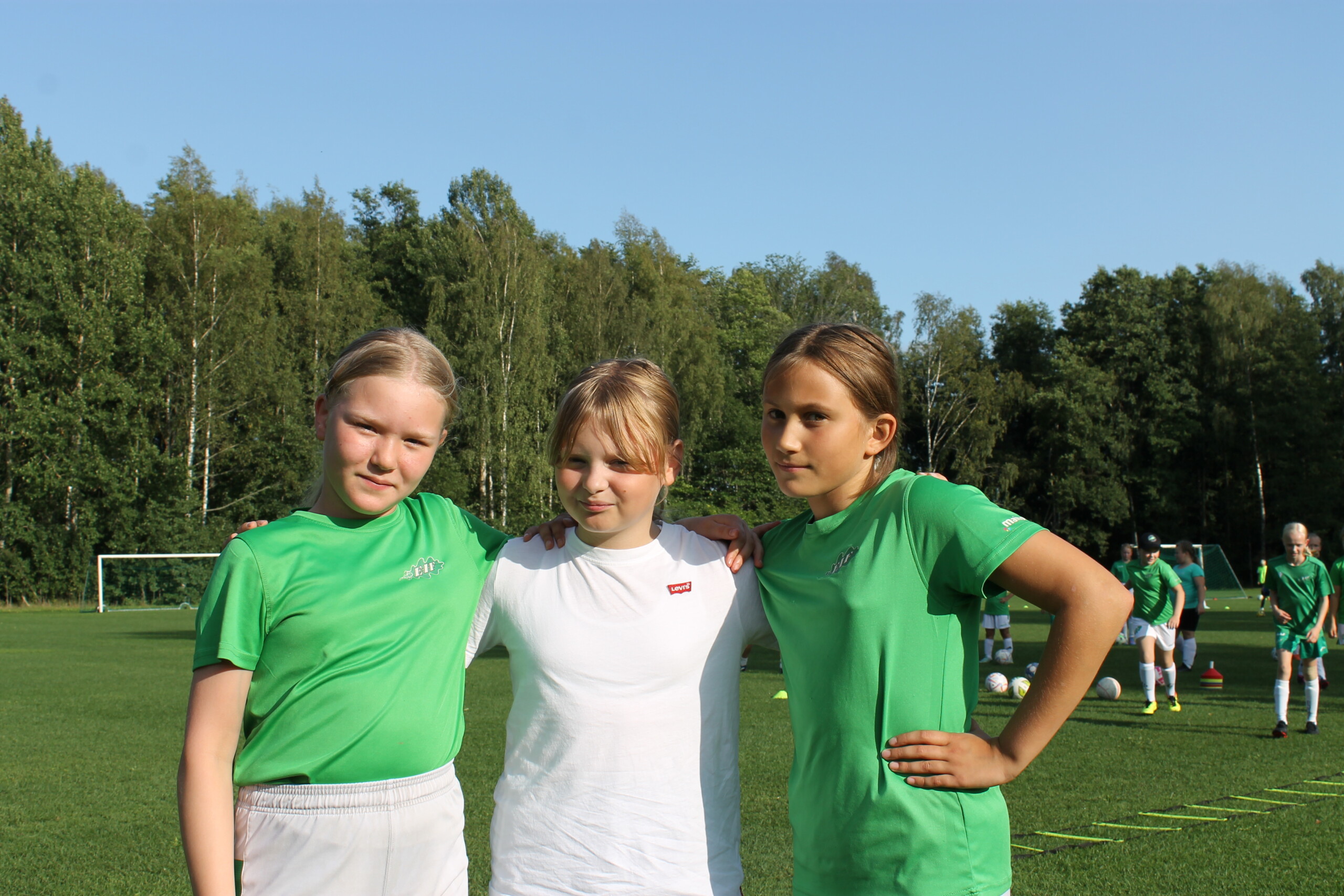 Tre flickor står bredvid varandra på en fotbollsplan och ler.