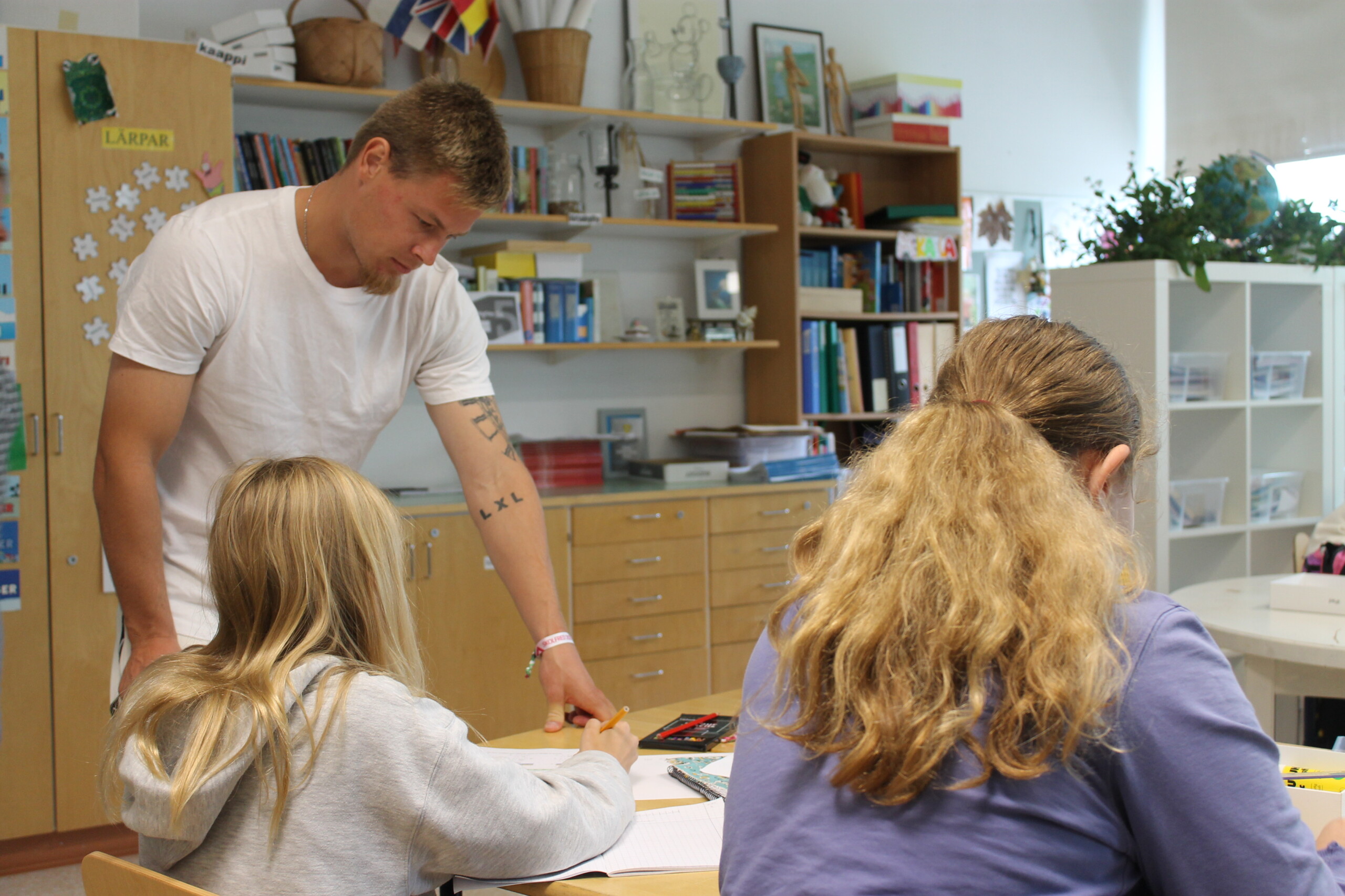 En ung kille hjälper elever i årskurs 5B i Seminarieskolan i Ekenäs. På bilden ser man honom hjälpa två elever i ett klassrum, eleverna sitter med ryggarna mot kameran. Mannen är en fotbollsspelare i EIF.