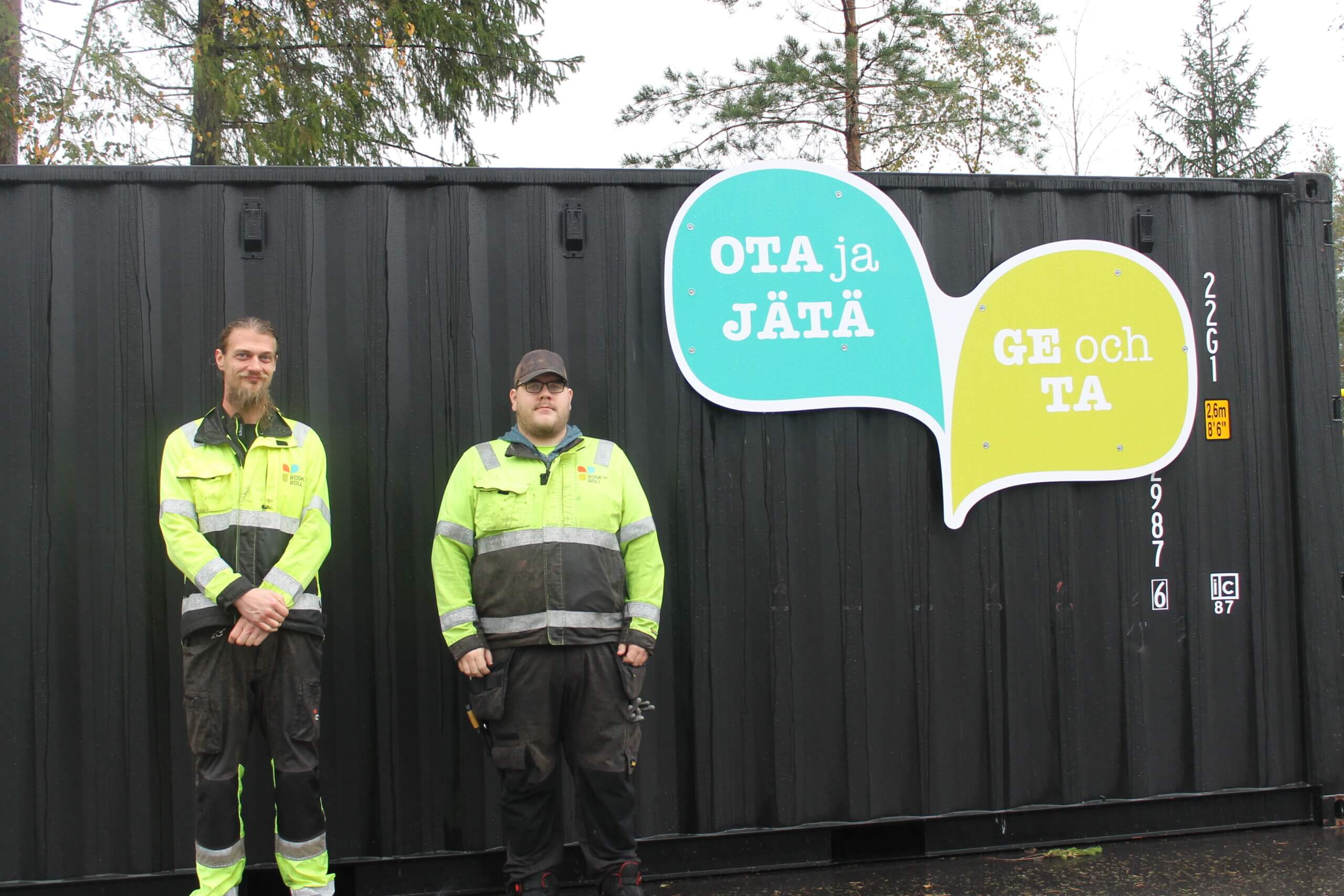 Två unga män står bredvid varandra klädda i reflexkläder. Bakom dem finns en svart container. Till höger om männen syns en skylt som säger "ge och ta". Männen arbetar på rosknroll i Karis.